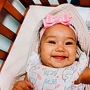 Zya is registered to the contest to win money with this photo: baby, smiling, infant, pink_bow, headband, unicorn_print, onesie, crib, wooden_crib, blanket, happy, cute, child, person, indoors, portrait, hand, skin, face, lying_down