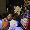 man, dog, resting, tattoo, beard, white_tank_top, brown_dog, indoor, decor, sculpture, pillow, bed, shelf, mirror, box, electronics, cigarette, tattooed_arm, curious_dog, relaxation