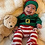 baby, infant, smiling, elf_costume, striped_pants, green_top, teddy_bear, soft_toy, blanket, fluffy, cute, happy, holiday, festive, child, portrait, indoors, cozy, playful, lying_down