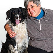 Mina participe au concours pour gagner de l'argent avec cette photo : woman, person, dog, border_collie, black_and_white, fluffy, long_hair, tongue_out, smiling, portrait, companion_animal, pet, cuddling, seated, gray_sweater, blue_shirt, black_pants, white_background, indoor, happy