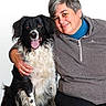 woman, person, dog, border_collie, black_and_white, fluffy, long_hair, tongue_out, smiling, portrait, companion_animal, pet, cuddling, seated, gray_sweater, blue_shirt, black_pants, white_background, indoor, happy
