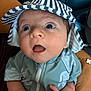 baby, infant, child, hat, striped_hat, blue_outfit, open_mouth, wide_eyes, hand, adult_hand, bracelet, curious, expression, close_up, portrait, person, indoor, cute, soft_lighting, holding