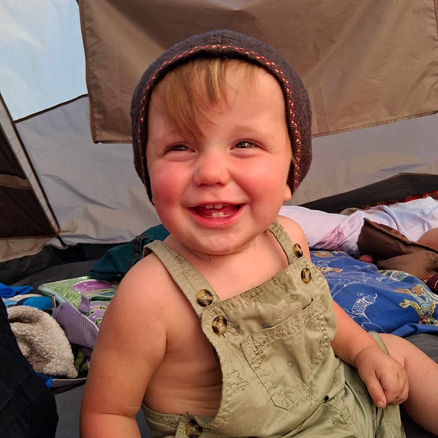 Derek is registered to the contest to win money with this photo: baby, blanket, camping, child, clothing, cute, daylight, fabric, happy, hat, nature, outdoor, overalls, person, playful, portrait, seated, smiling, tent, toddler