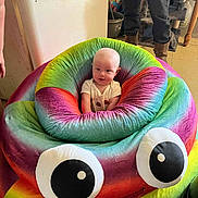 Malachi is registered to the contest to win money with this photo: baby, child, plush, rainbow, colorful, indoor, furniture, funny, smiling, person, seat, toy, googly_eyes, cozy, happy, cushion, casual_clothing, floor, adult, legs