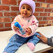 Aurelia joined the competition — help win amazing prizes! baby, child, infant, beanie, pink_sweater, hearts, blue_bottle, tongue_out, brick_wall, countertop, hand, sitting, cute, casual_clothing, indoor, person, smiling, playful, denim_pants, sock