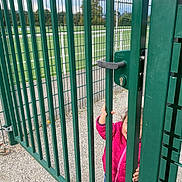 Loujayne participe au concours pour gagner de l'argent avec cette photo : child, clothing, curious, daylight, fence, gate, goalposts, gravel, green, handle, jacket, metal_bars, nature, outdoor, person, pink, playground, sports_field, toddler, trees