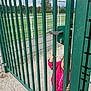 child, clothing, curious, daylight, fence, gate, goalposts, gravel, green, handle, jacket, metal_bars, nature, outdoor, person, pink, playground, sports_field, toddler, trees