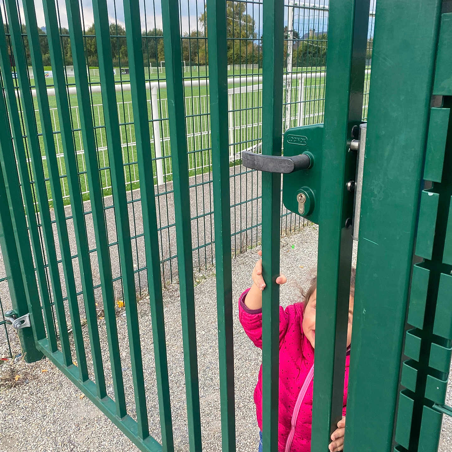 Loujayne participe au concours pour gagner de l'argent avec cette photo : child, clothing, curious, daylight, fence, gate, goalposts, gravel, green, handle, jacket, metal_bars, nature, outdoor, person, pink, playground, sports_field, toddler, trees