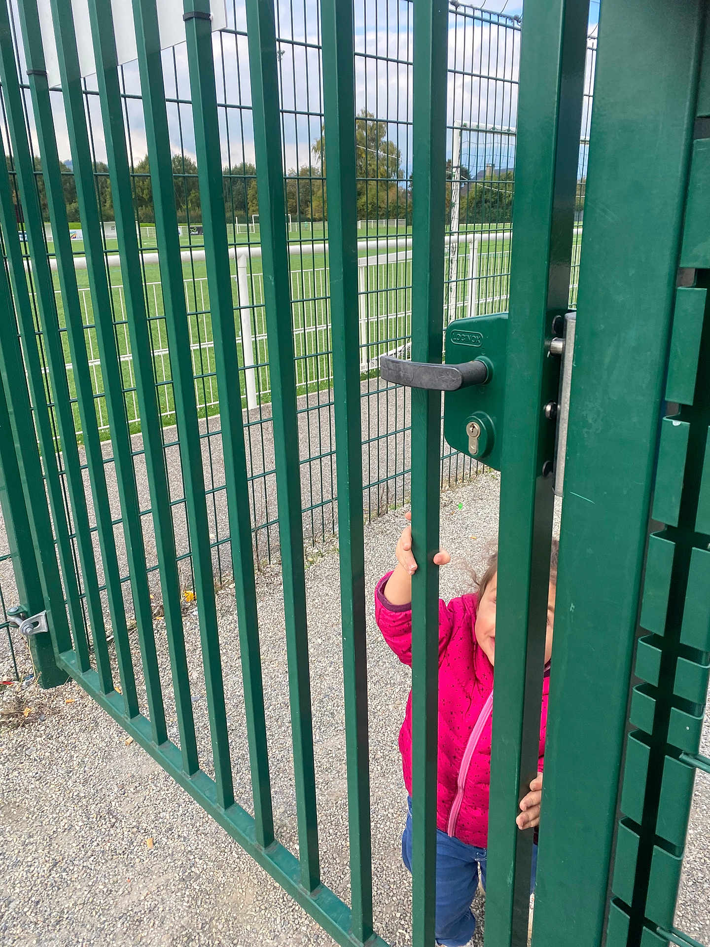 Loujayne participe au concours pour gagner de l'argent avec cette photo : child, clothing, curious, daylight, fence, gate, goalposts, gravel, green, handle, jacket, metal_bars, nature, outdoor, person, pink, playground, sports_field, toddler, trees