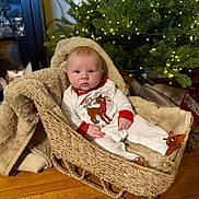 Henry is registered to the contest to win money with this photo: baby, basket, blanket, christmas_tree, clothing, cozy, decor, face, floor, footwear, furniture, holiday, indoors, lights, person, plush, reindeer, sitting, wood_floor, young_child