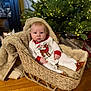 baby, basket, blanket, christmas_tree, clothing, cozy, decor, face, floor, footwear, furniture, holiday, indoors, lights, person, plush, reindeer, sitting, wood_floor, young_child