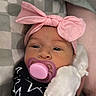 awake, baby, bed, black_outfit, blanket, checkered_blanket, child, close_up, cute, face, headband, indoor, infant, lying_down, mitten, pacifier, parent_arm, pink_bow, portrait, star_pattern