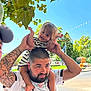 Mélody participe au concours pour gagner de l'argent avec cette photo : toddler, child, man, tattoo, shoulder_ride, outdoor, daylight, blue_sky, tree, leaves, street, crosswalk, flag, bunting, happy, playful, smile, striped_clothing, beard, person