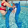 adult, blue_slide, child, family, fun, net_fence, outdoor, person, play, safety, shallow_water, smiling, splash, summer, sunlight, swimming_pool, swimsuit, water, water_park, water_slide