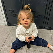 Mélody a rejoint le concours — aidez-le/la à gagner de superbes lots ! casual_clothing, child, curious, floor, flooring, gold_shoes, hair, indoor, keys, locker, looking_at_camera, navy_pants, person, ponytail, sitting, small_hands, tiles, toddler, white_sweater, young_child