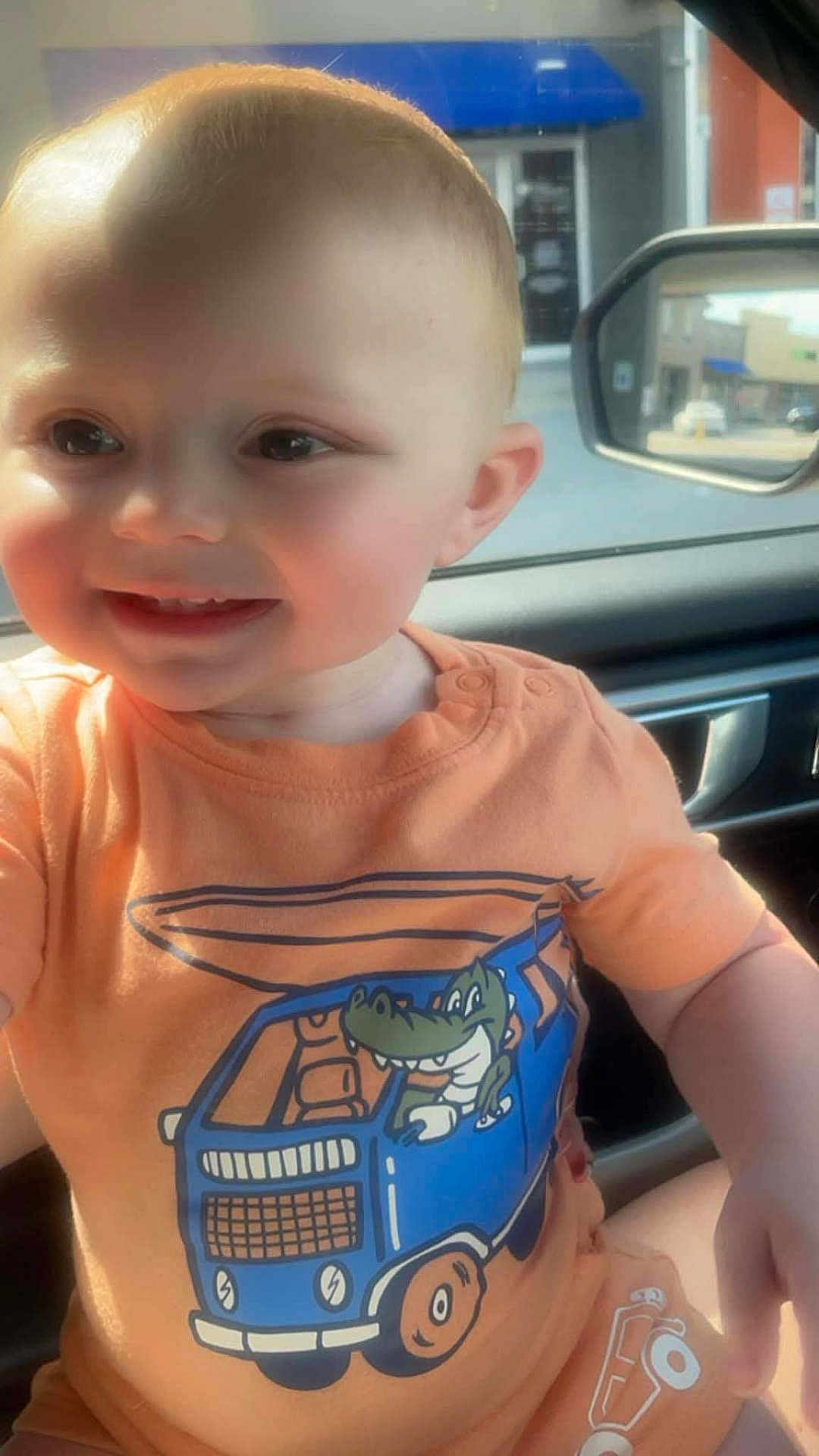 David is registered to the contest to win money with this photo: baby, child, smiling, orange_clothing, cartoon, crocodile, van, vehicle, car_interior, sunlight, portrait, cute, happy, infant, face, seat, window, daylight, young_child, casual
