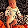 Leyann participe au concours pour gagner de l'argent avec cette photo : baby, infant, onesie, dinosaur_print, bedspread, orange_pillow, patterned_bedding, lying_down, serious_expression, human, child, cute, indoors, skin, legs, feet, hands, head, face, newborn