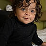 child, toddler, portrait, curly_hair, face, eyes, looking_at_camera, black_clothing, sitting, bed, bedroom, indoor, close_up, photography, serious_expression, cute, dark_clothing, brown_hair, soft_lighting, home