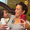 child, toddler, mother, parent, family, portrait, selfie, mirror, smiling, happy, indoors, home, baby, infant, white_clothing, gray_shirt, red_phone, tattoo, hair_bun, hearts