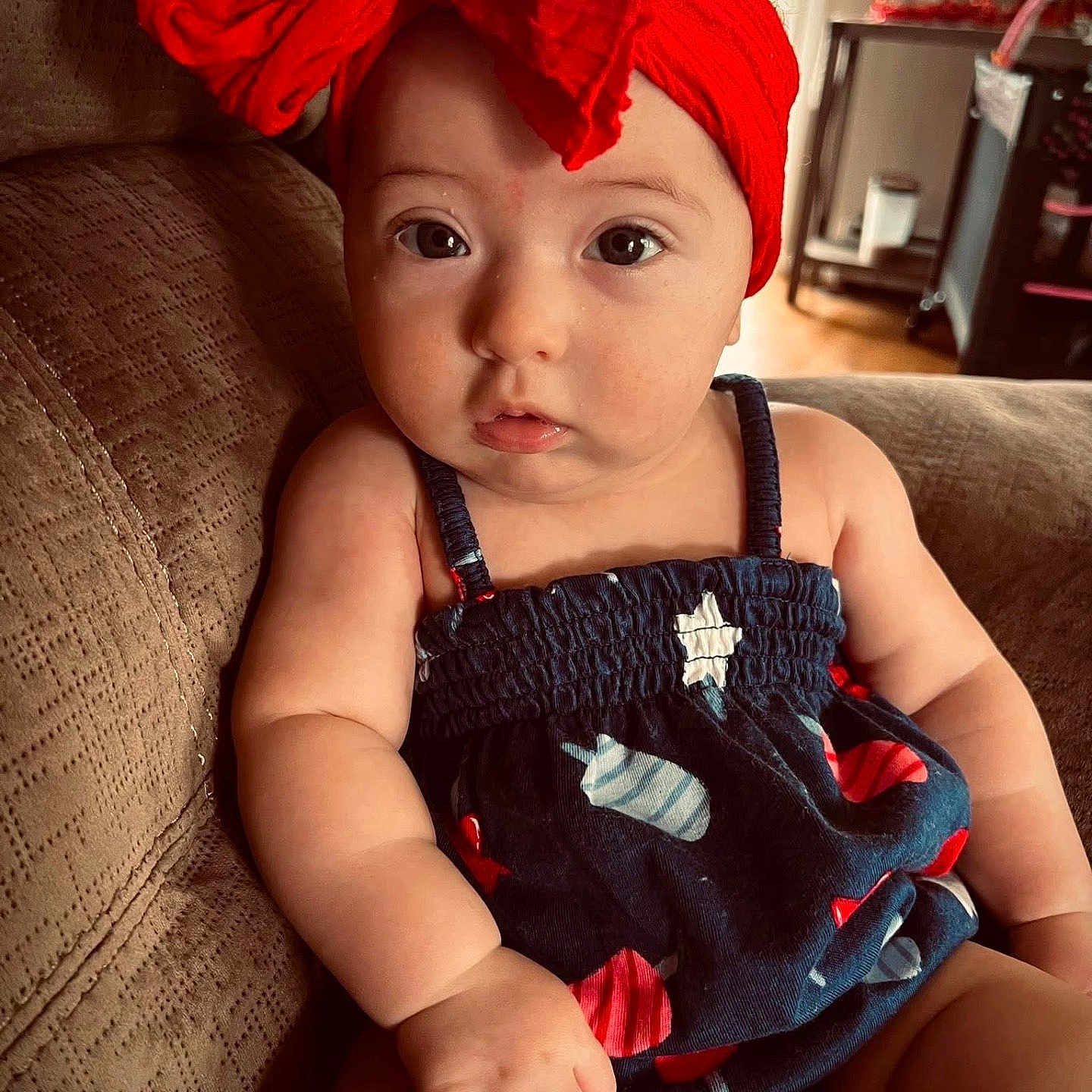 Kylie is registered to the contest to win money with this photo: arm, baby, child, clothing, couch, cute, expression, face, furniture, hand, headband, indoor, infant, living_room, patterned_outfit, person, portrait, red_bow, sitting, skin