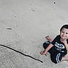 child, barefoot, concrete, stick, sitting, smiling, playful, casual_clothing, outdoor, young, person, short_hair, black_shirt, black_pants, barefoot_boy, expression, gesture, daylight, ground, posing