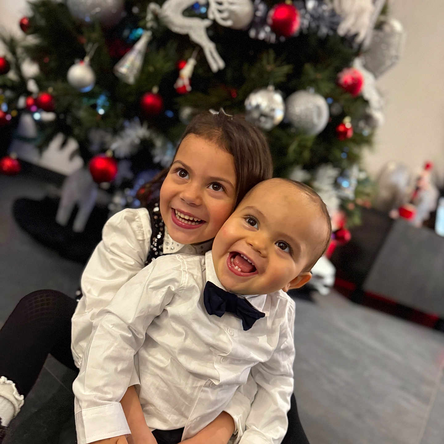 Sandro a rejoint le concours — aidez-le/la à gagner de superbes lots ! child, children, christmas_tree, holiday, smile, happy, embrace, festive, decorations, ornaments, white_shirt, bow_tie, black_pants, black_shoes, indoor, family, cute, joyful, toddler, sibling