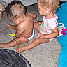 children, siblings, toddler, baby, sitting, carpet, toy_car, toy, playtime, indoors, curly_hair, diaper, pink_shirt, barefoot, family, play_area, blue_mat, infant, portrait, candid