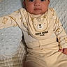 baby, infant, child, portrait, lying_down, sitting, indoors, bed, blanket, pillow, bear_pattern, bib, onesie, beige_clothing, cute, serious_expression, looking_at_camera, facial_portrait, home, soft_furnishings