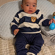 Leo is registered to the contest to win money with this photo: baby, infant, child, portrait, sitting, smiling, bed, pillow, cushion, toy, rattle, striped_clothing, navy_outfit, bear_design, sneakers, indoors, bedroom, cozy, happy, photography