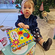 Léonye participe au concours pour gagner de l'argent avec cette photo : child, toddler, book, christmas_tree, ornament, gift_box, floor, indoor, holiday, toy, decor, person, dress, shoe, expression, face, hand, window, tile_floor, light