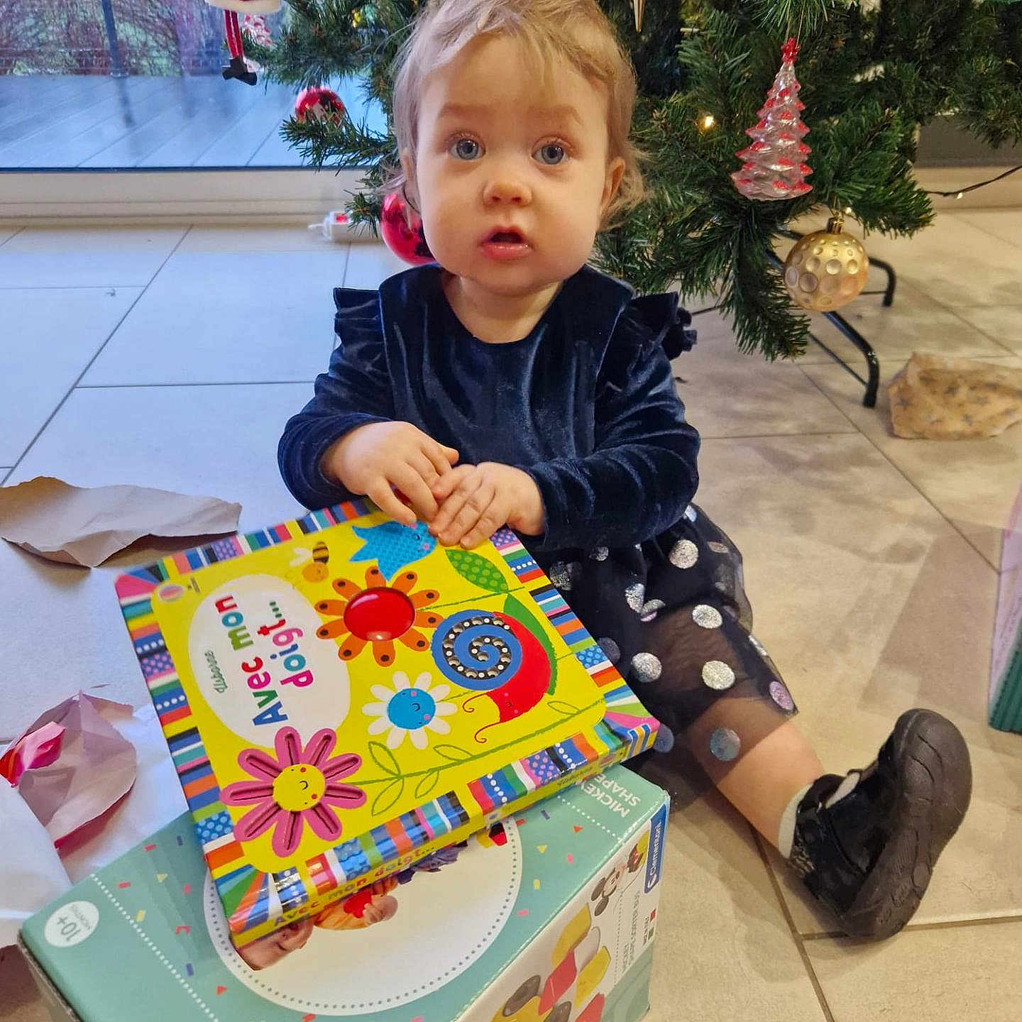 Léonye participe au concours pour gagner de l'argent avec cette photo : book, child, christmas_tree, decor, dress, expression, face, floor, gift_box, hand, holiday, indoor, light, ornament, person, shoe, tile_floor, toddler, toy, window