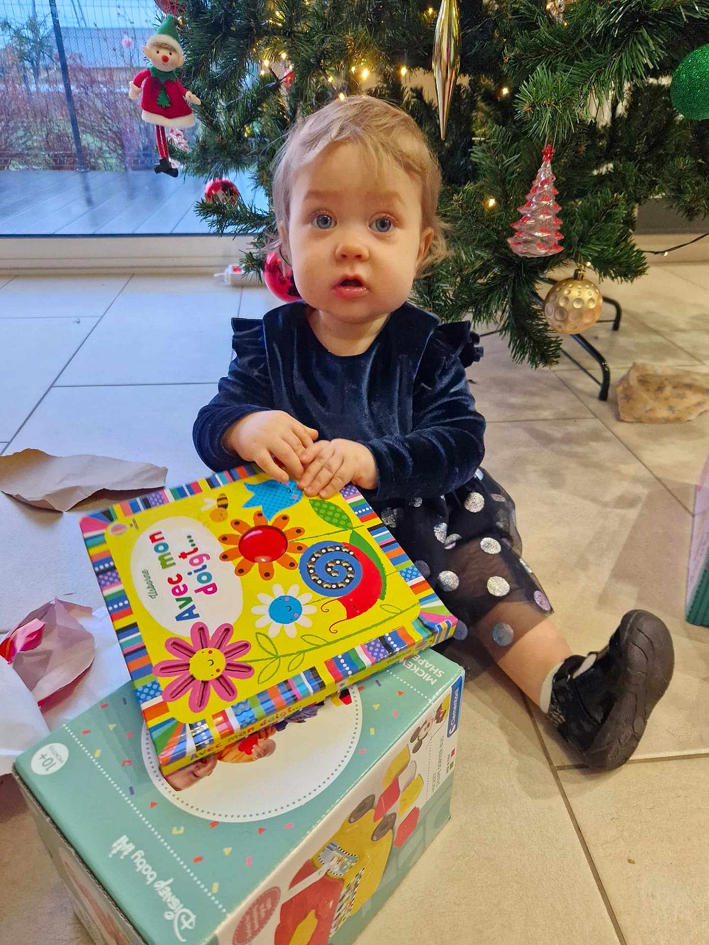 Léonye participe au concours pour gagner de l'argent avec cette photo : child, toddler, book, christmas_tree, ornament, gift_box, floor, indoor, holiday, toy, decor, person, dress, shoe, expression, face, hand, window, tile_floor, light
