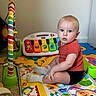 baby, child, sitting, portrait, blue_eyes, short_hair, red_shirt, black_shorts, socks, smiling, playroom, toy, musical_toy, toy_piano, play_mat, puzzle_mat, colorful, indoors, carpet, baby_photo