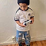 toddler, child, indoor, beanie, black_hat, white_shirt, adidas, ripped_jeans, blue_jeans, black_sneakers, game_controller, wooden_floor, wall, standing, looking_down, casual_clothing, floor_rug, necklace, pom_pom, young_child