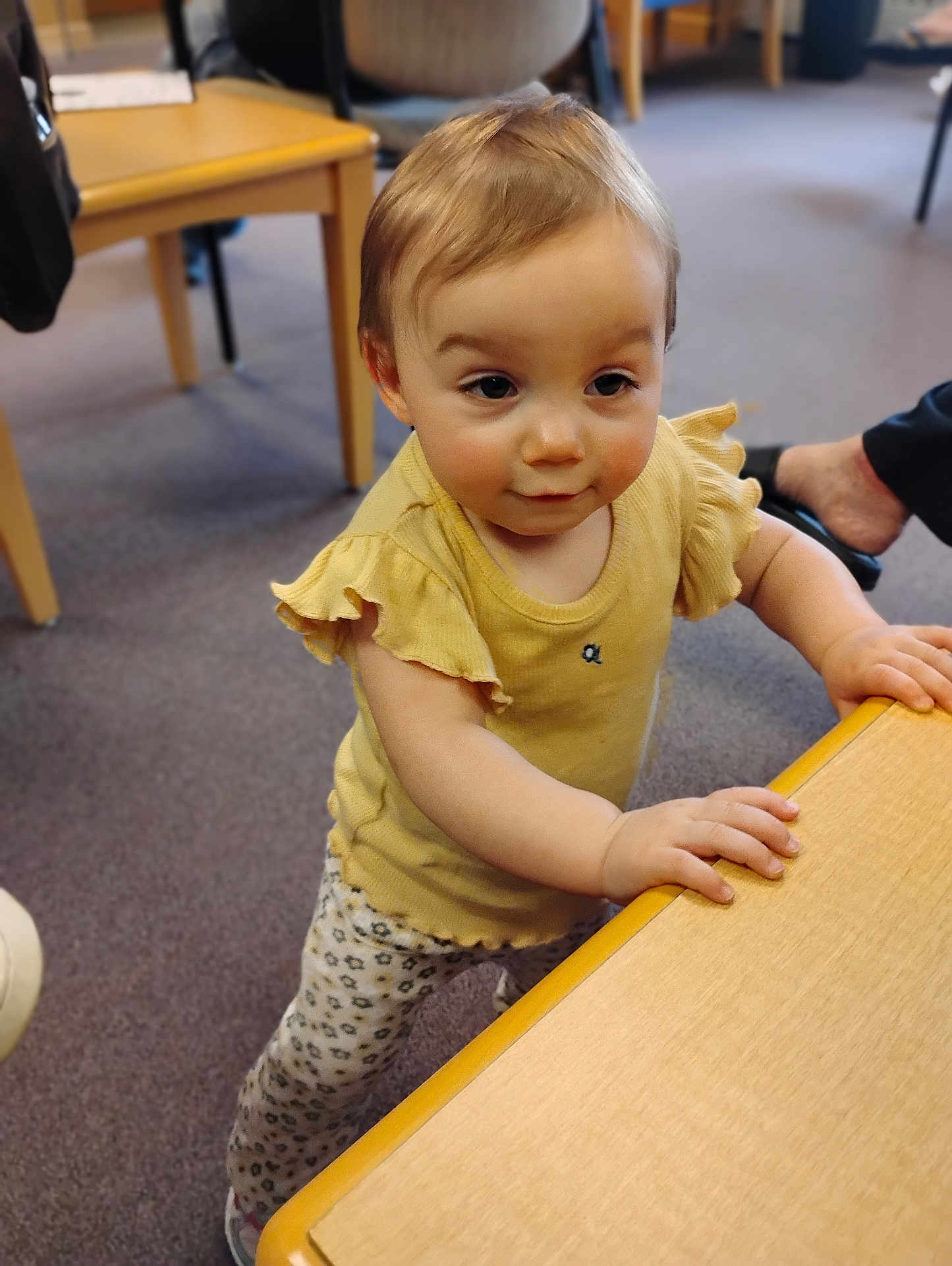 Amelia is registered to the contest to win money with this photo: toddler, child, indoor, yellow_top, patterned_pants, standing, table, carpet, curious, person, furniture, hand, face, short_hair, leaning, young_child, casual_clothing, floor, footwear, room