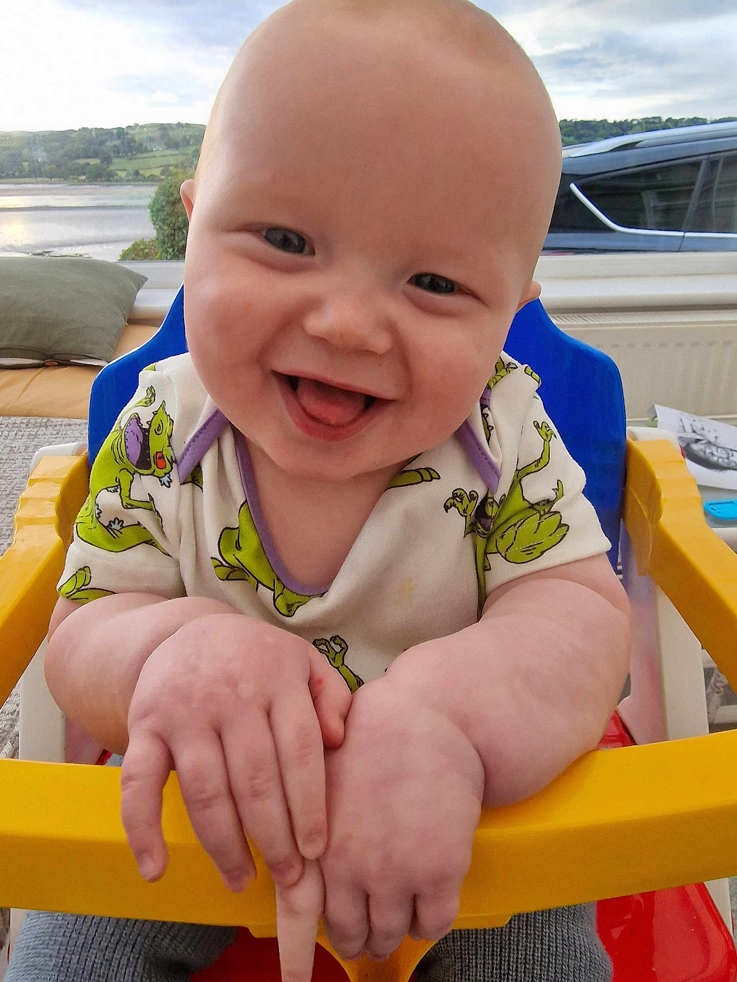 Mason is registered to the contest to win money with this photo: baby, smiling, high_chair, indoor, child, cute, happy, toddler, clothing, furniture, face, tongue_out, frog_pattern, window, scenic_view, hands, skin, person, seat, cheerful