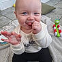 baby, child, tongue_out, smiling, indoor, blanket, toy, floor, cushion, tiled_floor, sitting, cute, happy, clothing, long_sleeve, pants, hand, face, playful, infant