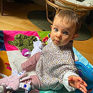 Madeline a rejoint le concours — aidez-le/la à gagner de superbes lots ! baby, child, play_mat, toy, indoor, floor, curious, blue_eyes, hand, pink_pants, patterned_shirt, plush_toy, pacifier, wooden_floor, chair, colorful, soft_toys, cushion, expression, person