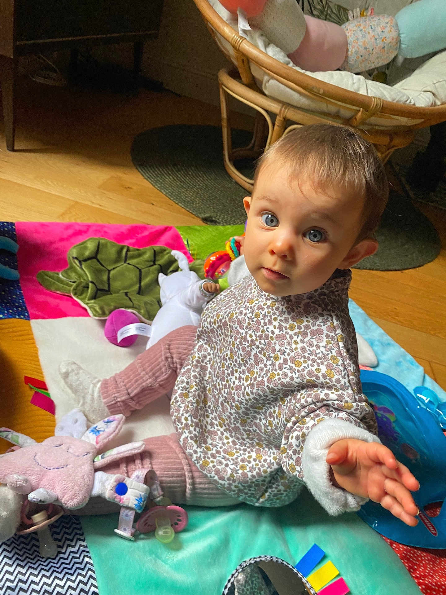 Madeline a rejoint le concours — aidez-le/la à gagner de superbes lots ! baby, child, play_mat, toy, indoor, floor, curious, blue_eyes, hand, pink_pants, patterned_shirt, plush_toy, pacifier, wooden_floor, chair, colorful, soft_toys, cushion, expression, person
