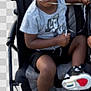 child, stroller, sneakers, grey_shirt, black_shorts, footwear, puzzled_expression, young_boy, sitting, outdoor, person, toy, plastic_cup, hand, curly_hair, casual_clothing, baby_stroller, legs, black_shoes, transparent_background