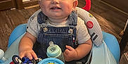 Saylor is registered to the contest to win money with this photo: baby, walker, toy, motorcycle, bottle, smiling, child, indoor, wooden_floor, denim_overalls, shirt, face, happy, playtime, furniture, door, wall_art, floor, seat, cute