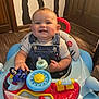 baby, walker, toy, motorcycle, bottle, smiling, child, indoor, wooden_floor, denim_overalls, shirt, face, happy, playtime, furniture, door, wall_art, floor, seat, cute