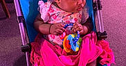 Amirah is registered to the contest to win money with this photo: baby, stroller, toy, blanket, infant, child, seated, carpet, indoor, clothing, foot, hand, curly_hair, pink, blue, wooden_chair, play, focus, young, cute