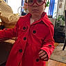 toddler, child, red_coat, heart_shaped_glasses, braided_hair, beads, bows, indoor, wooden_floor, curtains, furniture, plant, portrait, fashion, serious_expression, home_decor, natural_light, window, dress, person
