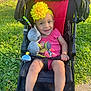 baby, child, stroller, toy, headband, yellow_flower, pink_clothing, smile, happy, outdoor, grass, sunlight, footwear, sitting, infant, playful, cute, person, daylight, park
