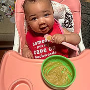 Blue Dream joined the competition — help win amazing prizes! baby, high_chair, bib, spoon, food, messy_face, pink, green_bowl, feeding, infant, cute, smiling, indoor, child, person, seat, hair, expression, table, meal