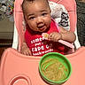 baby, high_chair, bib, spoon, food, messy_face, pink, green_bowl, feeding, infant, cute, smiling, indoor, child, person, seat, hair, expression, table, meal