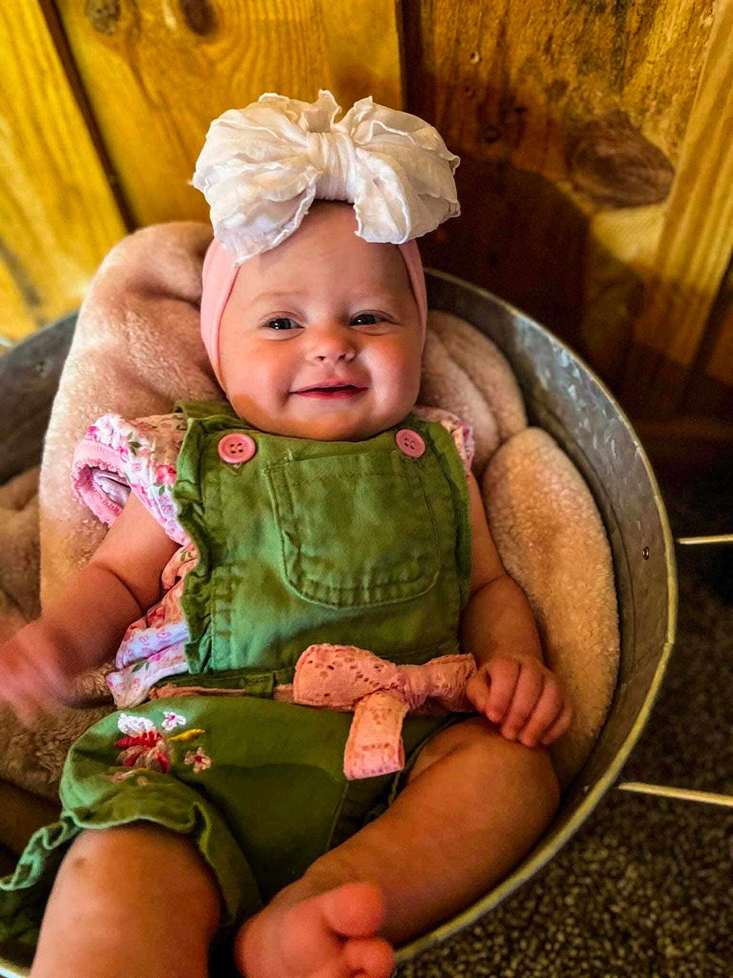 Piper is registered to the contest to win money with this photo: baby, infant, smiling, headband, bow, green_overalls, pink_bow, blanket, basket, wooden_background, cute, child, portrait, happy, indoor, soft_texture, comfortable, clothing, person, skin