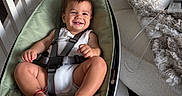 Enzo is registered to the contest to win money with this photo: baby, smile, baby_rocker, harness, toy, hanging_toys, indoor, chair, blanket, furniture, floor, wood_floor, person, infant, happy, clothing, foot, hand, head, face
