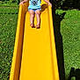 child, slide, playground, yellow_slide, grass, outdoor, summer, blonde_hair, blue_shirt, denim_shorts, sunlight, trees, nature, playing, fun, person, barefoot, shadow, daylight, park